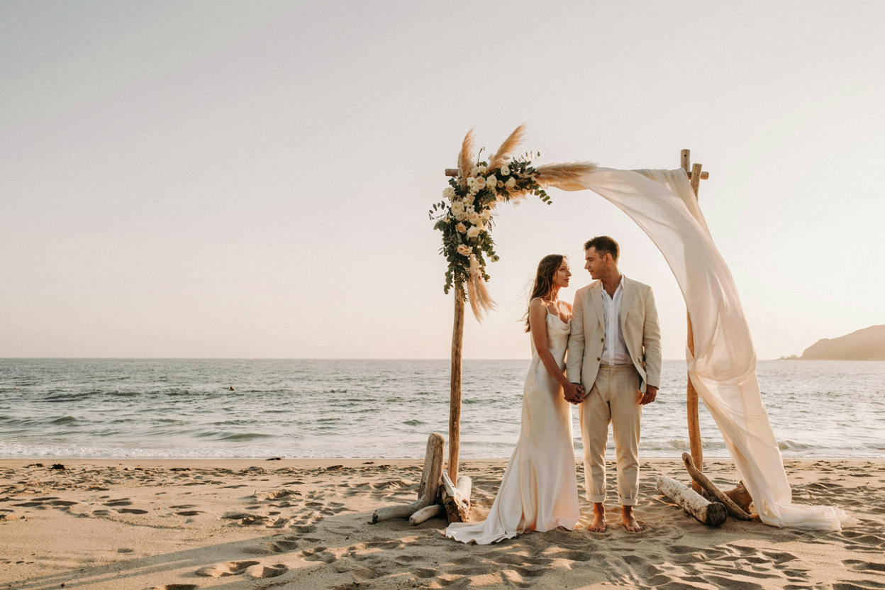 A couple standing beneath a floral arch on a quiet beach at sunset, representing the Sacred Union Ceremony for two partners.